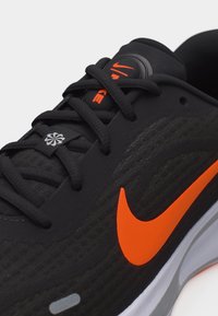 Črni tekaški čevelj Nike z oranžnim logotipom, črnimi vezalkami in vidnim logotipom na jezičku na belem ozadju.