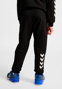 Sorte træningsbukser med konisk snit, tekstureret stof og hvide chevron-detaljer på den nederste del af benet, båret med blå sportssko.