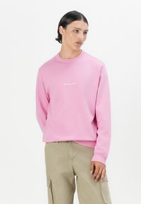 Sweat-shirt léger rose avec un col ribbed et des poignets, comportant un petit détail texte blanc sur la poitrine. Associé à un pantalon cargo kaki.