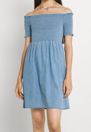 Woman wearing a blue off-shoulder smocked dress with short sleeves and a heart-shaped necklace, standing against a plain background.