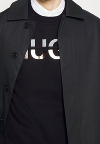 Cappotto nero sopra una felpa nera con scritte riflettenti in argento e oro "HUG". Tessuto liscio con bottoni sul cappotto.