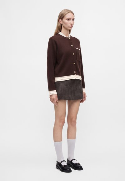Femme portant un cardigan marron boutonné avec une bordure en dentelle blanche, une mini-jupe à fines rayures, des chaussettes blanches montantes et des chaussures Mary Jane noires, debout.