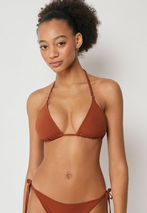 Jeune femme aux cheveux bouclés portant un haut de bikini à col halter couleur rouille et un bas assorti sur un fond clair uni.