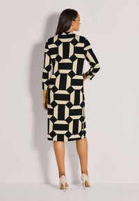 Eine Frau steht mit dem Rücken zur Kamera und trägt ein knieumspielendes Kleid mit geometrischem Muster in Schwarz und Beige, mit 3/4-Ärmeln und beigen High Heels.