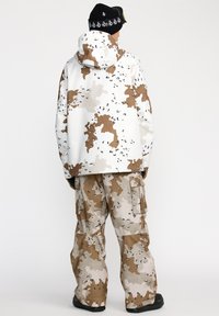 Witte en bruine camouflage-gepatroniseerde buitenkleding met een capuchon, gecombineerd met bijpassende cargobroeken. De stof lijkt waterdicht en heeft een gladde textuur.