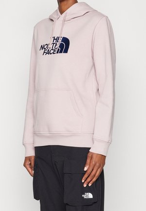 Osoba ubrana w jasnoróżową bluzę z kapturem The North Face oraz czarne spodnie cargo z przednią kieszenią, stojąca na jednolitym tle.