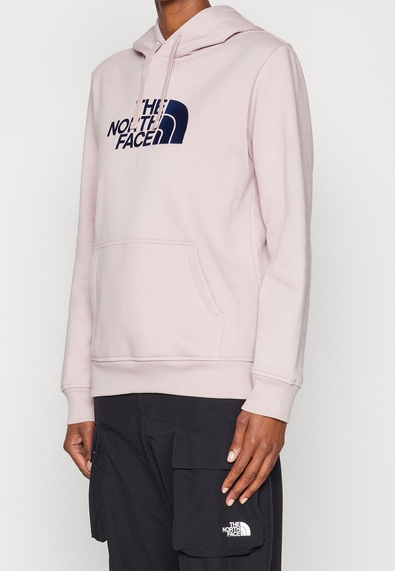 Personne portant un hoodie The North Face rose clair et un pantalon cargo noir avec une poche avant, debout devant un fond uni.