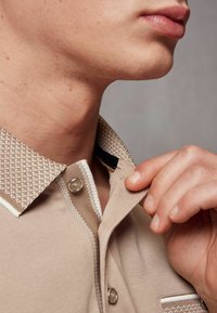 Beige-paita, jossa on tekstuurilla varustettu kaulus, nappisuljenta ja korostetut raidat. Läheltä otettu kuva kaulasta ja kädestä, joka säätää kaulusta. Pehmeä kangas.