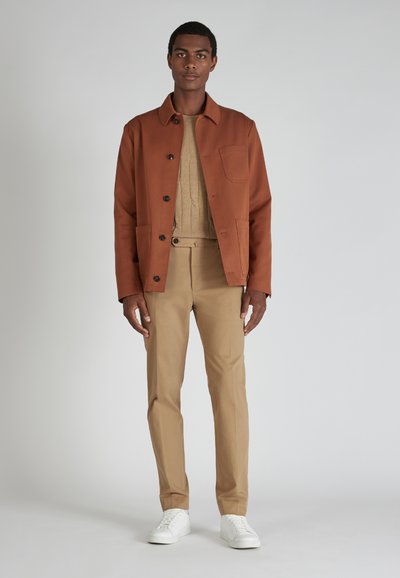 Braune Baumwolljacke mit schwarzen Knöpfen und Tasche, beigefarbener Strickpullover, beiger Hose und weißen Sneakers. Das Modell steht vor einem hellen Hintergrund.