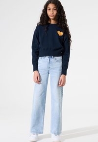 Pull bleu marine avec un détail en forme de cœur jaune ; jean léger à coupe ample bleu clair ; baskets blanches ; mannequin se tient devant un fond neutre.