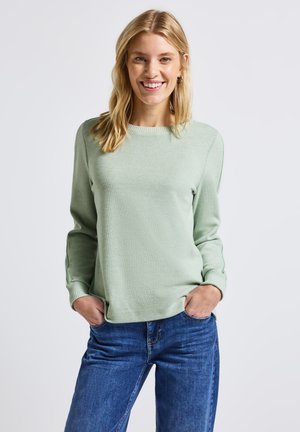 Blondynka uśmiechająca się, ubrana w jasnozielony sweter z długim rękawem i niebieskie dżinsy, z rękami w kieszeniach, stojąca na jednolitym jasnym tle.