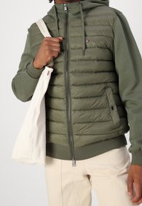 Pufferjakke med hætte i olivengrøn, med lynlås foran, ribstrikkede manchetter, diagonalt quiltet og med et broderet logo.