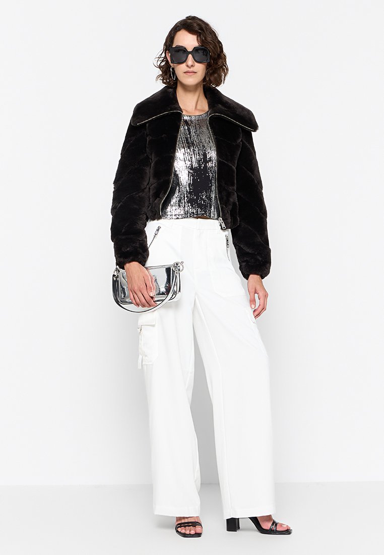 Schwarze Kunstpelz-Cropped-Jacke, metallisch silbernes Oberteil, weiße weit geschnittene Hose und silberne Handtasche. Model trägt dunkle Sonnenbrille und Riemchensandalen.