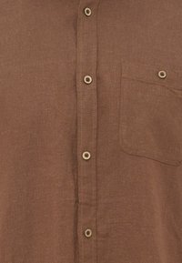 Τμήμα καφέ λινής πουκάμισου που δείχνει τα μπροστινά κουμπιά και μια κουμπωμένη τσέπη στο στήθος στη δεξιά πλευρά.