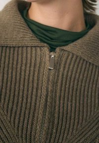 Hnědý pletený kardigán s vertikálním žebrovaným vzorem, zapínáním na zip a širokým límcem přes tmavě zelený oděv pod ním.