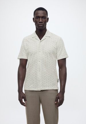 Mann trägt ein weißes kurzärmliges Hemd mit Knöpfen und Lochmuster sowie hellbraune Hose, steht vor einem einfarbigen Hintergrund.