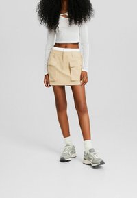 Jupe cargo beige avec plis devant et grandes poches latérales. Associée à un haut blanc à manches longues côtelé et des baskets grises.