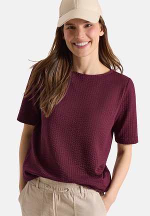 Femme souriante portant une casquette beige, un haut bordeaux à manches courtes texturé et un pantalon beige, les mains dans les poches sur un fond uni.