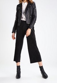 Černá kožená motorkářská bunda se stříbrnými zipy, bílá blůza s puntíky a černé plisované široké kalhoty s kratší délkou, doplněné černými kotníkovými botami.