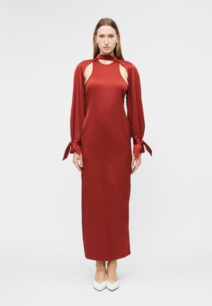 Γυναίκα που στέκεται, φοράει ένα μακρύ κόκκινο φόρεμα με ανοιχτά κοψίματα στους ώμους, μακριά μανίκια δεμένα και λευκά μυτερά παπούτσια, μπροστά σε ένα απλό ανοιχτόχρωμο φόντο.