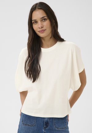 Femme aux longs cheveux foncés portant un haut blanc uni à manches courtes et un jean bleu, debout devant un fond neutre.