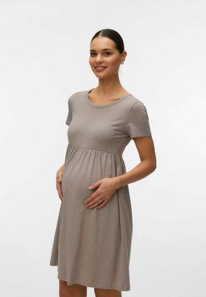 Pregnant woman smiling, wearing short-sleeve taupe maternity dress, hands gently resting on baby bump against a plain white background.