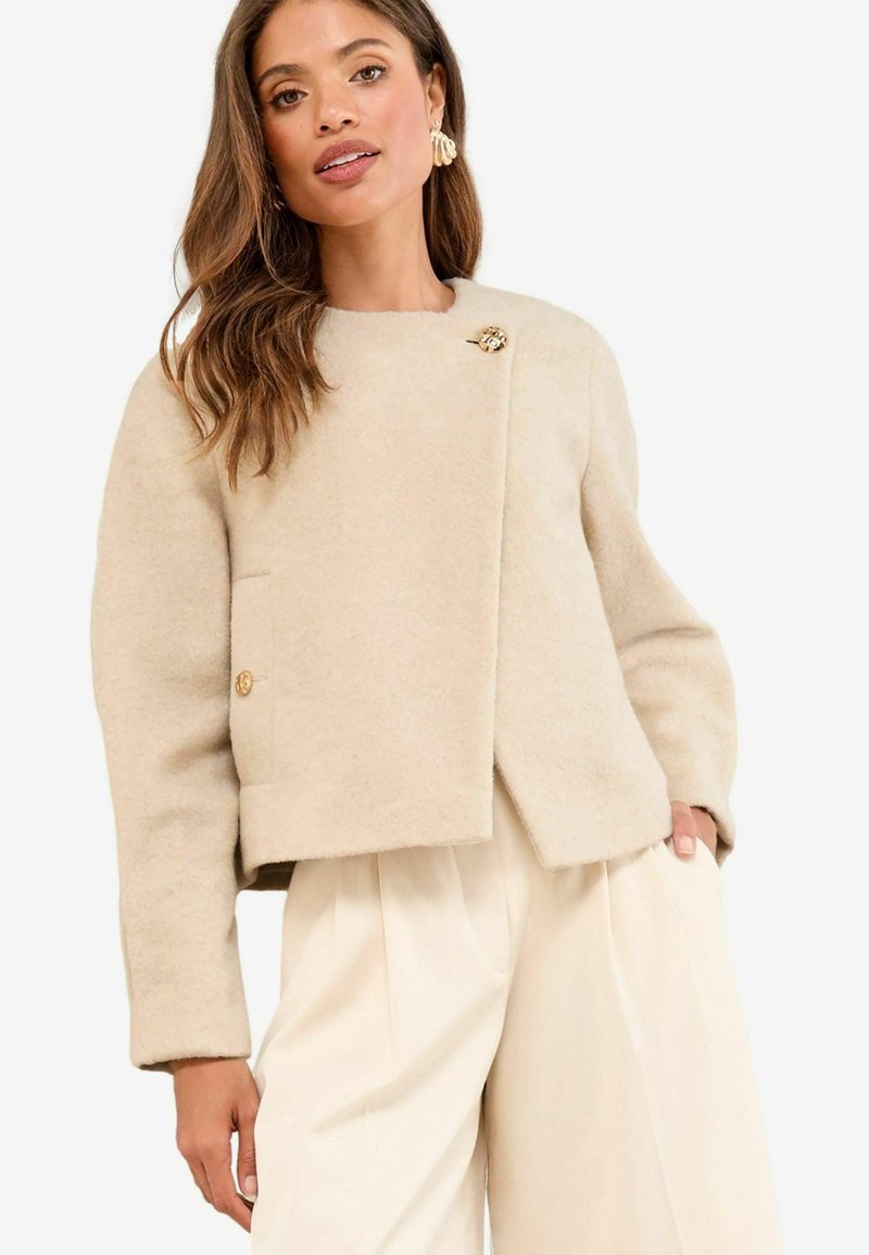 Vrouw in een beigekleurige wollen cropped jasje met gouden knopen en bijpassende wijde broek, poserend met één hand in de zak.