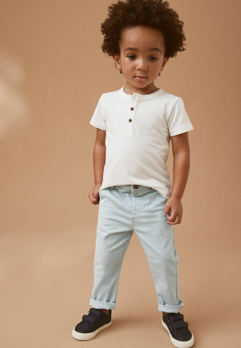 Weißes Kurzarm-Henley-Shirt mit Holzknöpfen, hellblaue aufgerollte Jeans und dunkle Sneakers. Vor einem beigen Hintergrund.