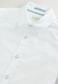 Weißes Hemd mit Kragen aus strukturiertem Stoff und markanten Knöpfen mit der Aufschrift "Baker by Ted Baker", flach auf einer hellen Oberfläche liegend.