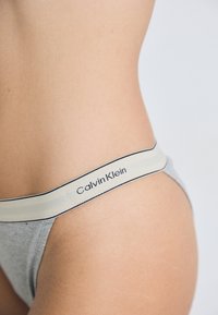 Close-up van het onderste deel van een persoon zijn torso die grijze Calvin Klein ondergoed draagt met een beige tailleband en zwarte tekst.