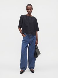 Femme portant un haut noir ample et un jean bleu large, tenant un sac noir d’une main dans la poche, debout devant un fond uni.