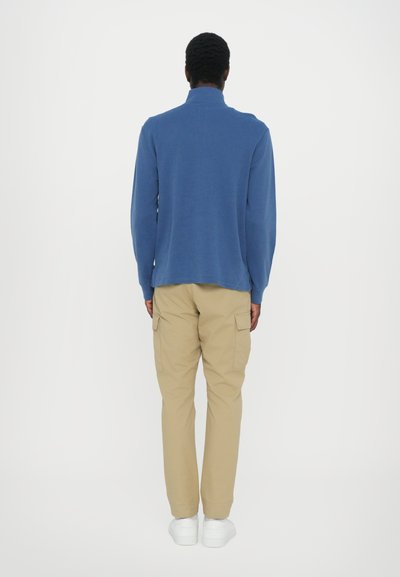 Blauwe lange mouwen overhemd met een staande kraag, gecombineerd met beige cargobroek en witte sneakers. De achterkant laat de stofstructuur en pasvorm zien.