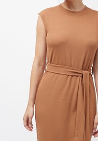 Brun ærmeløs kjole i et blødt, tekstureret strikket stof. Har en bæltet talje for en tætsiddende pasform. Simpelt, elegant design med en rund halsudskæring.