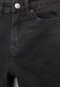 Detailní záběr na černé džíny zepředu, ukazující knoflík, oblast zipu a švy kapes.