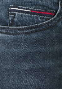 Mörkblå jeans i denim som visar bakfickan med en liten randig tygdetalj i marinblått, vitt och rött längs fickkanten.