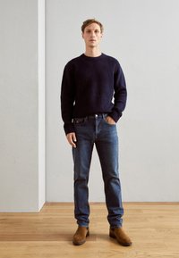 Marineblauwe gebreide trui, blauwe jeans en bruine suède laarzen. De outfit heeft een klassiek ontwerp met een gestructureerd breisel en een ontspannen pasvorm.