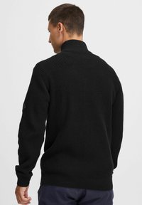 Suéter de punto negro con un patrón texturizado, cuello alto y puños acanalados. La parte trasera presenta un ajuste relajado y un diseño simple.