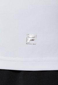 Vit träningströja med en liten silverlogotyp vid nederkanten, gjord av mjukt, fukttransporterande tyg med en figurnära design.