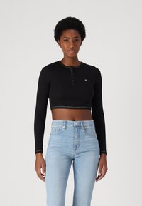 Sort crop top med lange ærmer, knaplukning ved halsen og kontrastsyet. Sammen med lyseblå højtaljede jeans.
