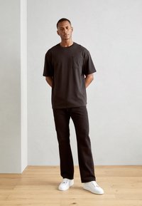 Mørkebrun kortærmet t-shirt med brystlomme, parret med sorte lige bukser og hvide sneakers på et trægulv.