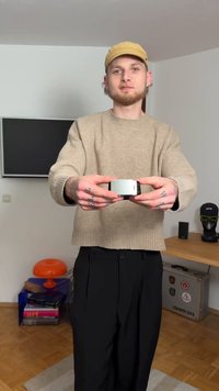 A compact camera with a silver front and black detailing, held by a person wearing a beige sweater and black pants, with tattoos visible.