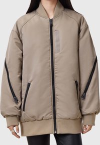 Khaki Bomberjacke mit glatter Textur, Reißverschlusstaschen und elastischen Bündchen. Verfügt über einen gerippten Kragen und ein dezentes Stoffpatch-Detail.