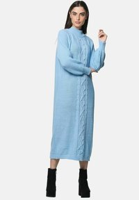 Kvinde iført en lyseblå midi sweaterkjole med kabelstrik og lange ærmer samt sorte plateau-ankelstøvler, stående mod en ensfarvet baggrund.