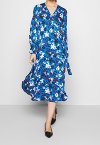 Blaues Blumenkleid mit langen Ärmeln, V-Ausschnitt und ausgestelltem Saum. Das Design umfasst weiße, gelbe und dunkelrote Blumen auf einem blauen Hintergrund.