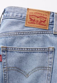 Jean en denim bleu clair montrant la poche arrière avec une couture orange et une étiquette en cuir marron Levi Strauss & Co. au-dessus de la poche.