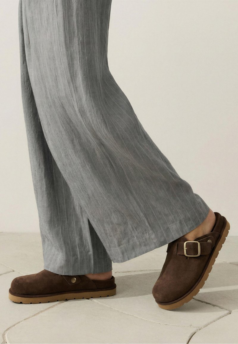 Sabots marron en daim sans lacets avec boucle métallique et semelles en caoutchouc clair, portés avec un pantalon ample gris à fines rayures sur un sol carrelé.