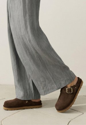 Saboturi maro din piele întoarsă, slip-on, cu cataramă metalică și tălpi din cauciuc deschis, purtate cu pantaloni largi gri cu dungi subțiri pe podea cu gresie.