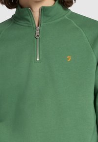 Grüner Fleecepullover mit hohem Kragen, ausgestattet mit einem silbernen Reißverschluss und kleinem orangefarbenem Logo auf der Brust. Glatte Textur und lockere Passform.