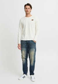 Langærmet cremefarvet skjorte med et lille broderet logo, parret med fadede blå jeans og sort-hvide stribede sneakers.