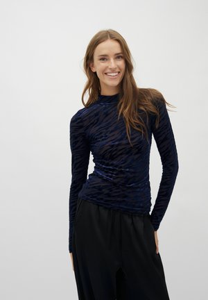 Nasmejana ženska z dolgimi svetlo rjavimi lasmi, oblečena v prosojno mornarsko modro bluzo z vzorcem in dolge rokave ter široke črne hlače, na enobarvnem ozadju.